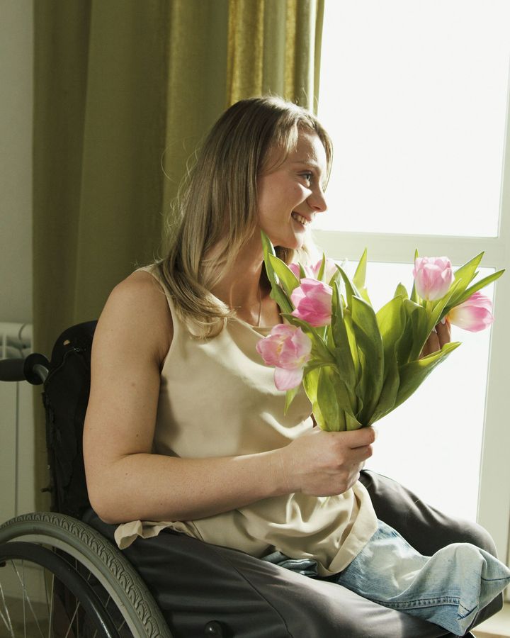 A smiling woman in a wheelchair holding pink tulips by a window.
